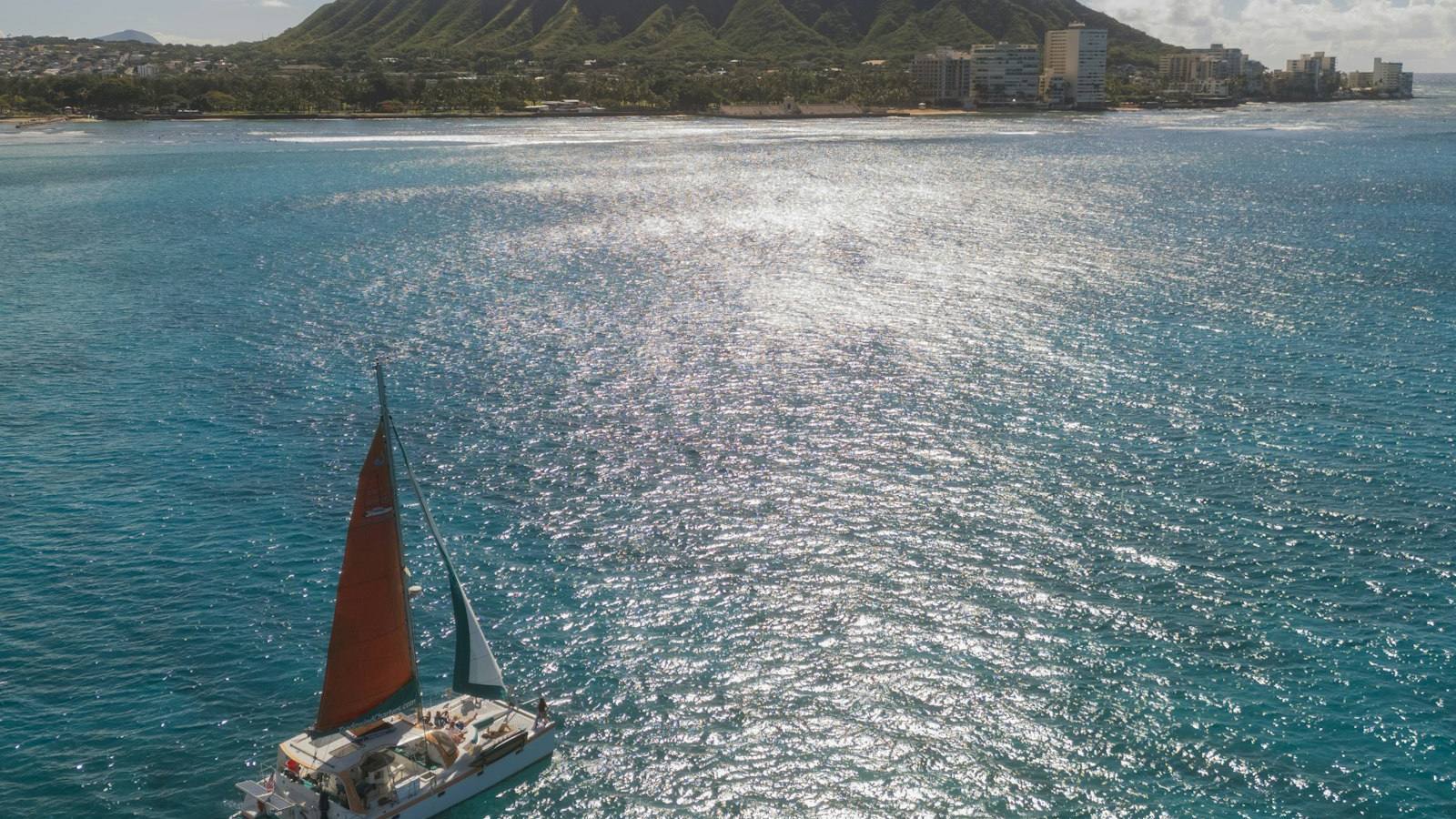 A catamaran under sail on the water off Waikiki, with Diamond Head and the shoreline behind it